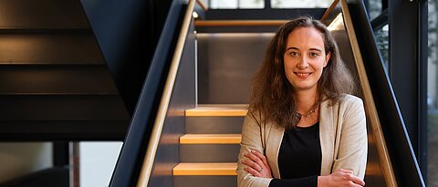 Eine Frau mit mittellangen dichten Haaren trägt einen hellen Blazer. Sie hält die Arme verschränkt. Sie steht vor einer Bibliothekstreppe